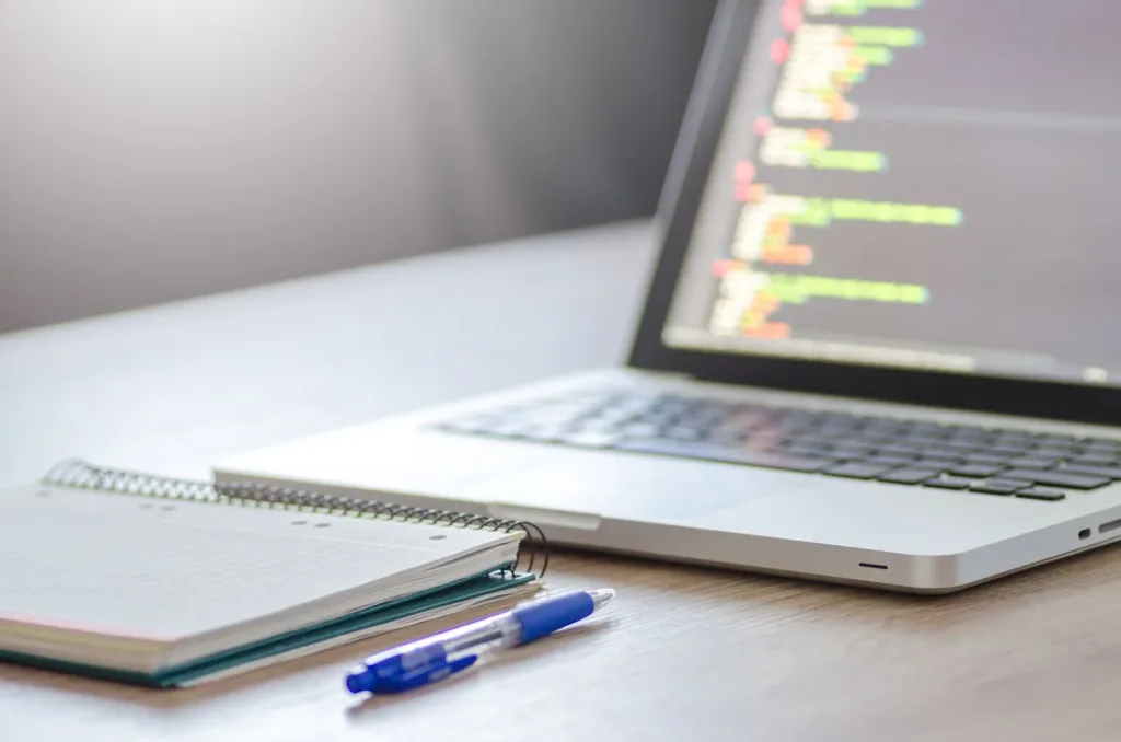 Laptop with code on screen beside a notebook and pen